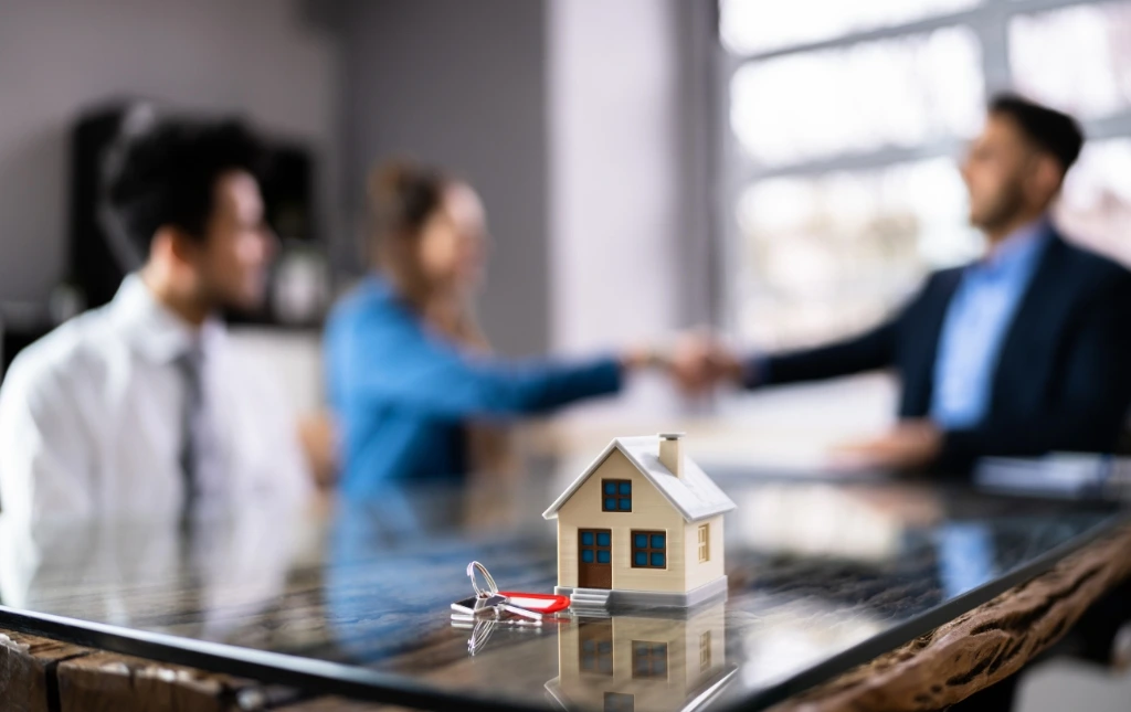 A-miniature-house-model-with-keys-sits-on-a-table-while-two-people-shake-hands-in-the-background-finalizing-a-real-estate-deal