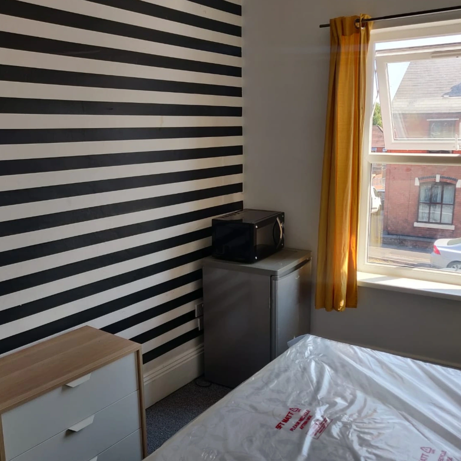 A-room-with-a-black-and-white-striped-wall-a-microwave-and-mini-fridge-and-a-window-with-yellow-curtains-overlooking-an-outside-street
