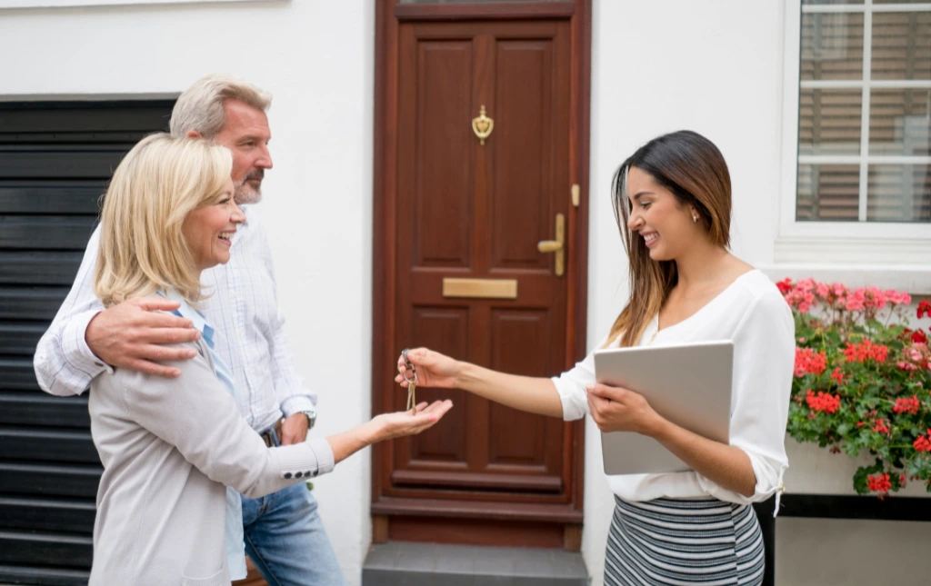 A-smiling-agent-hands-keys-to-a-happy-couple-in-front-of-a-new-home-representing-Moving-Beyond-Traditional-An-Alternative-to-Selling-Lettings