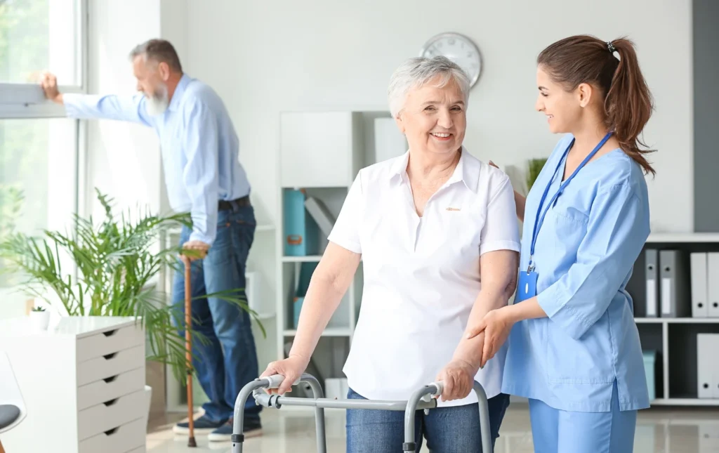 Caregiver-assisting-elderly-person-with-walker-in-residential-care-home-for-adults-with-fellow-resident-in-background