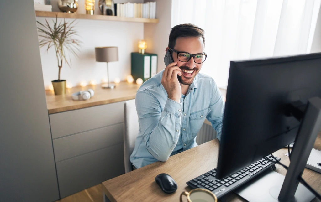 Smiling-man-in-glasses-talking-on-phone-while-working-on-desktop-computer